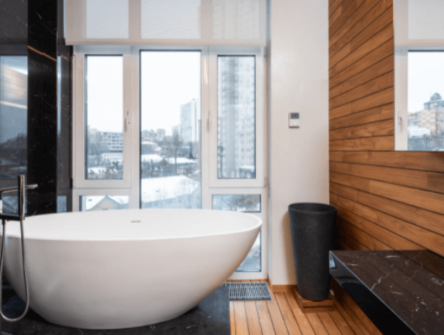 banheiro com piso e parede de madeira , uma banheira branca elevada do chão, pia preta do lado direito da imagem e uma janela na frente da banheira