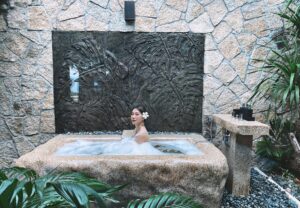 Mulher em uma banheira jacuzzi na área externa. A banheira é de pedras e há vegetação ao redor. capa da manutenção da Jacuzzi