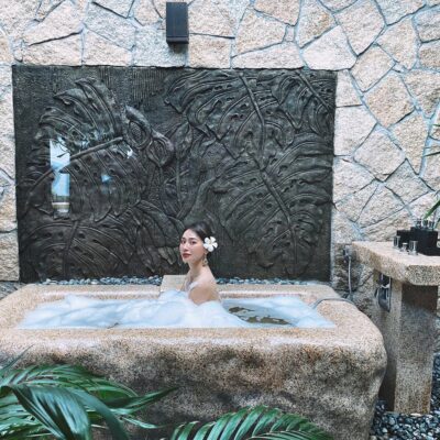 Mulher em uma banheira jacuzzi na área externa. A banheira é de pedras e há vegetação ao redor. capa da manutenção da Jacuzzi